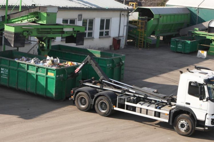 Biały samochód (kontenerowiec) wraz z zielonym kontenerem. W tle biały budynek