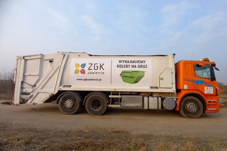 Samochód ciężarowy typu śmieciarka. Biała zabudowa i pomarańczowa kabina